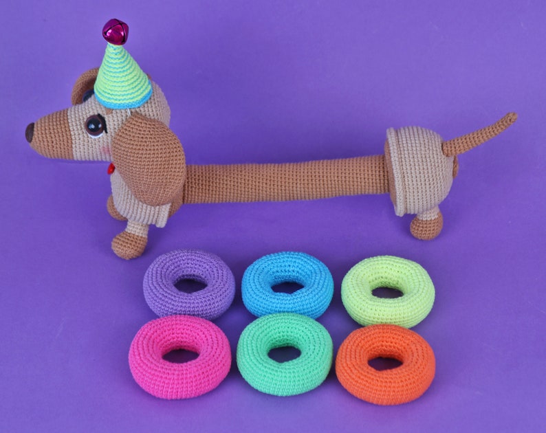 May include: A crocheted dachshund wearing a party hat and holding six colorful crocheted rings.