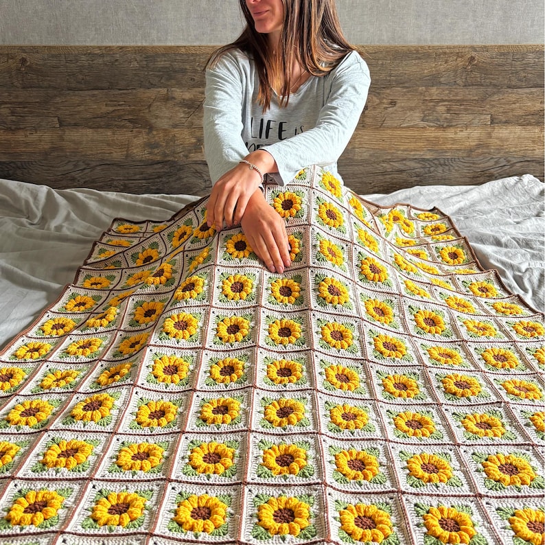 May include: A crocheted blanket with a sunflower pattern. The blanket is made of white, brown, and yellow yarn. The blanket is laid out on a bed.