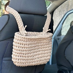 May include: A beige crocheted bag hanging from the headrest of a car seat. The bag has a woven texture and is made of thick yarn.