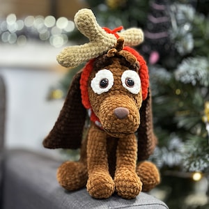 May include: A brown crocheted dog toy with a red and white crocheted reindeer antler headband. The toy has white eyes and a brown nose.