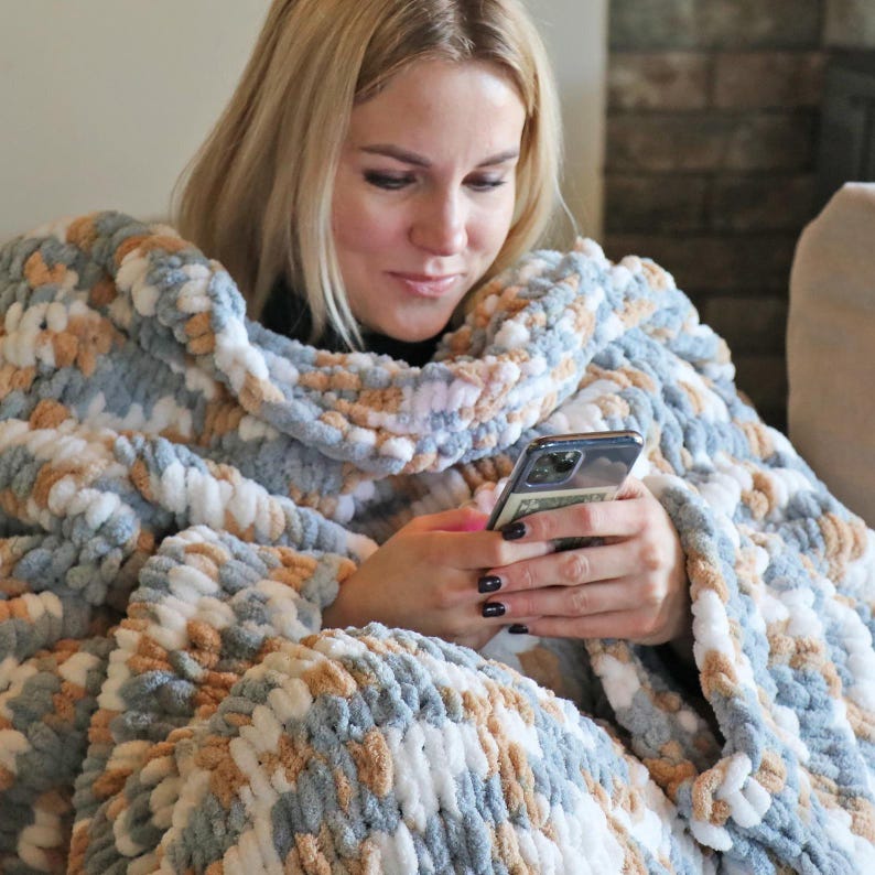 May include: A cozy, oversized blanket in shades of blue, white, and tan. The chunky knit blanket is wrapped around a person, providing warmth and comfort. The person is holding a smartphone.