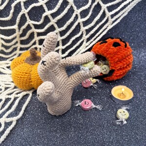 May include: A crocheted Halloween-themed display featuring a tan hand holding wrapped candies. An orange pumpkin-shaped candy holder and a small orange pumpkin are also present. A lit candle and a white crocheted spiderweb complete the scene.