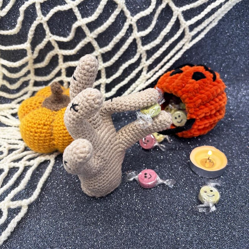 May include: A crocheted Halloween-themed display featuring a tan hand holding wrapped candies. An orange pumpkin-shaped candy holder and a small orange pumpkin are also present. A lit candle and a white crocheted spiderweb complete the scene.