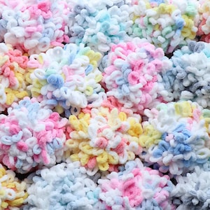 May include: A close-up shot of multiple fluffy yarn pom-poms in pastel shades. The colours include pink, blue, yellow, and white. The yarn has a soft, textured appearance, indicating a craft or DIY project, possibly for knitting or crochet.