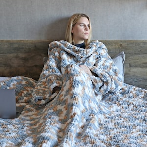 May include: A person is sitting on a bed wrapped in a large, soft, grey and white knitted blanket. The blanket has a fluffy texture and is made of yarn. The person is holding a laptop computer.