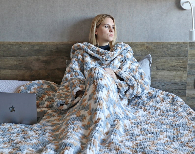 May include: A person is sitting on a bed wrapped in a large, soft, grey and white knitted blanket. The blanket has a fluffy texture and is made of yarn. The person is holding a laptop computer.