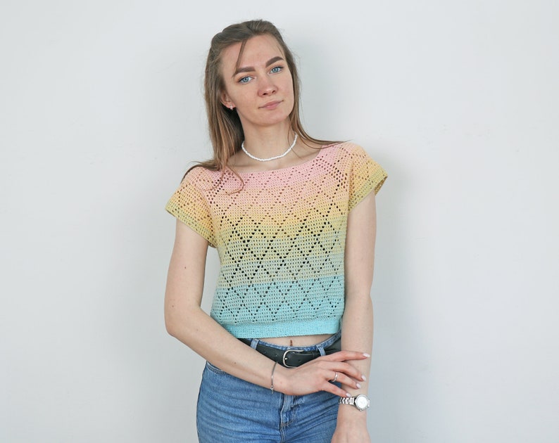May include: A pastel-colored crochet top with a diamond pattern. The top transitions from pink to yellow to blue. The short-sleeved shirt is paired with blue jeans and a black belt. A white necklace and watch are also visible.