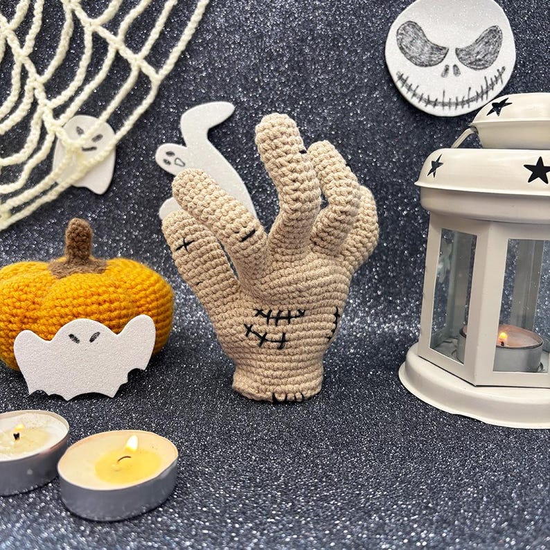 May include: A beige crocheted hand with black stitched details, surrounded by Halloween decorations. Includes a crocheted pumpkin, a white lantern, and a Jack Skellington-themed decoration. The scene is set with a spiderweb and ghost cutouts.