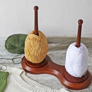 Puede incluir: Un soporte de madera para hilo con dos carretes de hilo. Un carrete es blanco y el otro es de color amarillo claro. El soporte de hilo está sobre una superficie blanca.