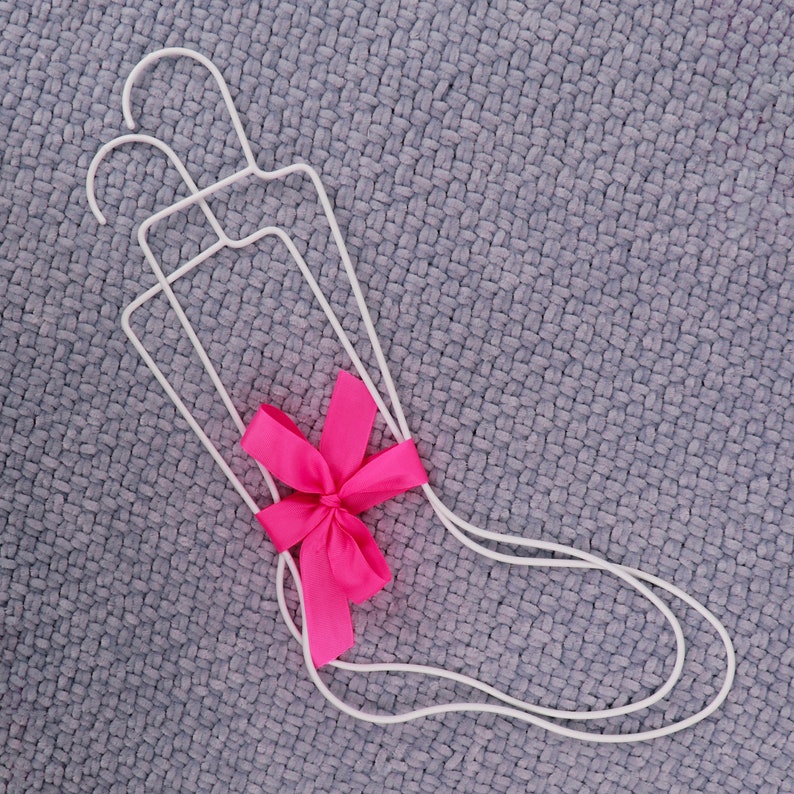 May include: Three white wire hangers are tied together with a pink bow. The hangers are on a gray textured surface.