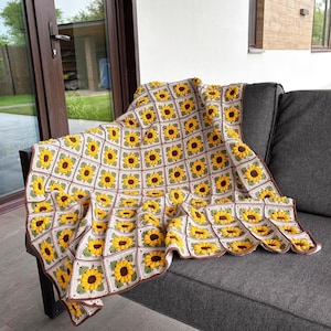 K&ouml;nnte beinhalten: Handgefertigte H&auml;keldecke mit einem sich wiederholenden Sonnenblumenmuster. Die Decke besteht aus cremefarbenem, gelbem, braunem und gr&uuml;nem Garn. Die Decke ist &uuml;ber ein graues Sofa drapiert und zeigt das komplizierte florale Design.