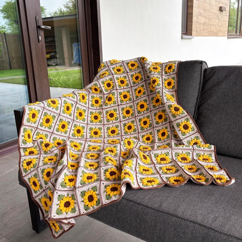 K&ouml;nnte beinhalten: Handgefertigte H&auml;keldecke mit einem sich wiederholenden Sonnenblumenmuster. Die Decke besteht aus cremefarbenem, gelbem, braunem und gr&uuml;nem Garn. Die Decke ist &uuml;ber ein graues Sofa drapiert und zeigt das komplizierte florale Design.