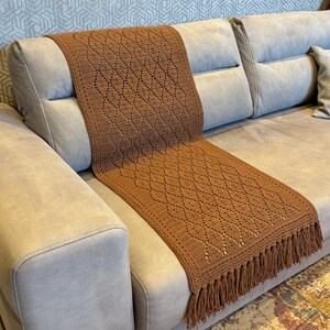 Puede incluir: Un cubre sofá de crochet marrón con un patrón de diamante y flecos. El cubre sofá está drapeado sobre un sofá marrón claro.