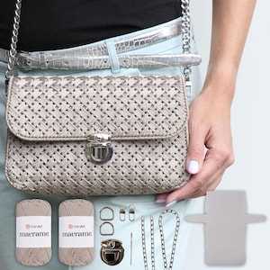 Może przedstawiać: Beżowa torebka z makramą z srebrnym zapięciem i łańcuszkowym paskiem. Torebka jest wykonana z dwóch szpul beżowej przędzy, srebrnego łańcuszka, dwóch srebrnych kółek D, dwóch srebrnych zatrzasków, srebrnej igły i beżowej podszewki.