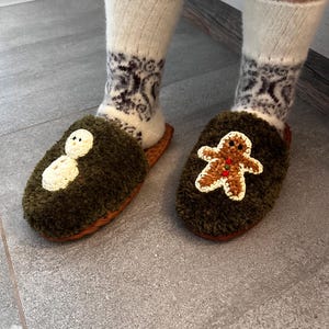 May include: A pair of green fuzzy slippers with embroidered holiday designs. One slipper features a snowman and the other features a gingerbread person.