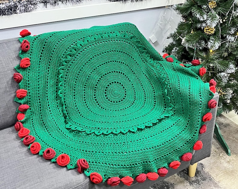 May include: A green crocheted round blanket with red pom-poms around the edge. The blanket is on a gray couch.