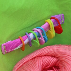 May include: A close-up of a green fabric bag with a pink and white striped strap. The strap has a row of colorful plastic stitch markers attached to it. A ball of pink yarn is visible in the bottom of the bag.