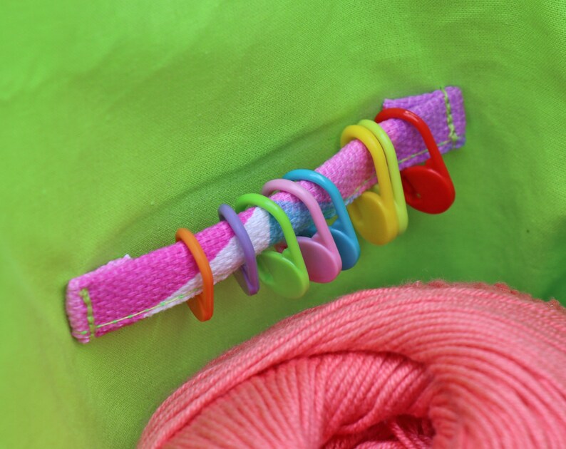 May include: A close-up of a green fabric bag with a pink and white striped strap. The strap has a row of colorful plastic stitch markers attached to it. A ball of pink yarn is visible in the bottom of the bag.