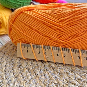 Puede incluir: Una gran madeja de hilo naranja con una regla de madera colocada delante. La regla mide en cent&iacute;metros y tiene hebras de hilo naranja colocadas encima. Se ven otras madejas de hilo en amarillo, verde y rosa en el fondo.