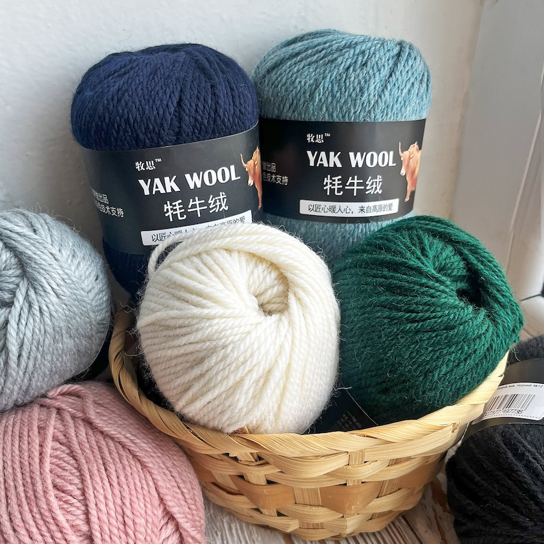 May include: A wicker basket filled with balls of yarn in various colors, including navy blue, light blue, white, green, pink, and gray. The yarn balls have labels in Chinese characters that read "YAK WOOL" and "牦牛绒", which translates to "Yak Wool".