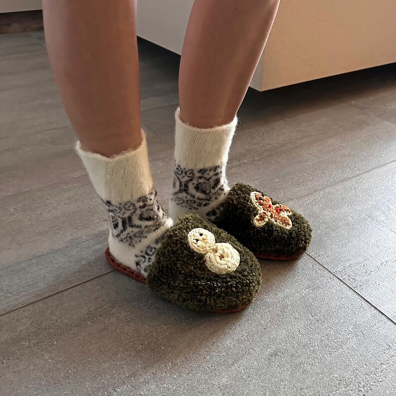 May include: A pair of green fuzzy slippers with embroidered snowman and gingerbread man designs. The slippers are worn with white socks with a black and white geometric pattern.