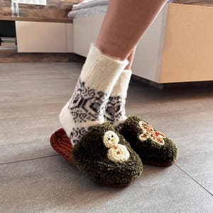 May include: A pair of green fuzzy slippers with embroidered snowman and gingerbread man designs. The slippers are worn with white socks with a black and brown geometric pattern.