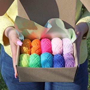 May include: A box filled with 12 balls of yarn in various colours, including pink, purple, blue, green, yellow, and orange. The yarn is arranged in rows in the box.