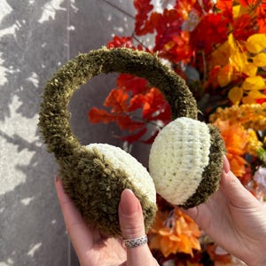 May include: Handmade green and cream-colored earmuffs. The ear covers are a combination of cream and green, while the headband is a fluffy green material. The earmuffs are designed to keep ears warm in cold weather. The background features fall foliage.