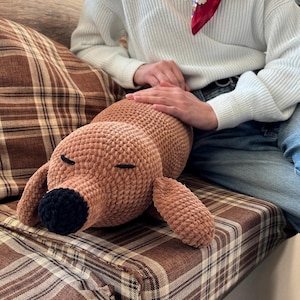 Könnte beinhalten: Ein braunes, gehäkeltes Plüschtier in Form eines Dackels mit schwarzen Augen und einer schwarzen Nase. Das Spielzeug liegt auf einem karierten Sofa.