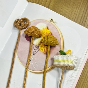 May include: Crocheted bookmarks in the shape of a pretzel, croissants, and a slice of cake. The bookmarks are brown, yellow, and white, with green and yellow accents. The bookmarks are on a book page with an embroidery hoop in the background.