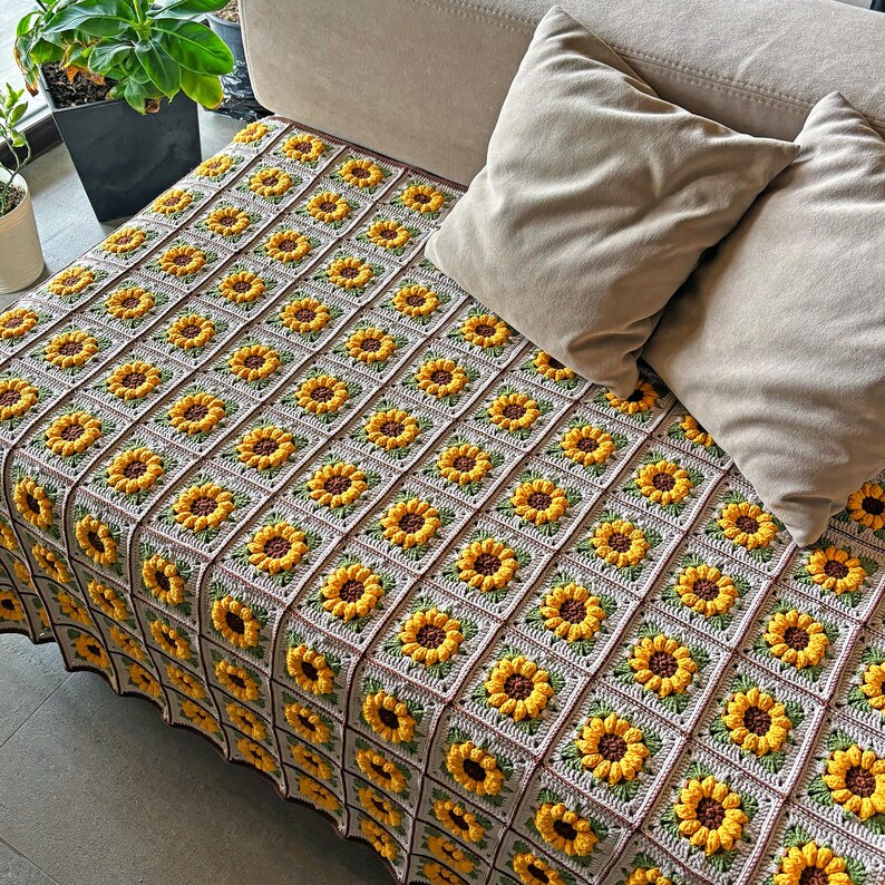 May include: A handmade crochet blanket featuring a repeating sunflower pattern. The blanket is made of yellow, brown, and green yarn, with a neutral background. The blanket is draped over a beige couch, with two matching pillows.
