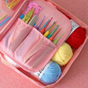 May include: Pink zippered pouch with multiple compartments for storing crochet hooks and yarn. The pouch is open and shows a variety of colourful crochet hooks and balls of yarn in red, yellow, and blue.
