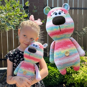 May include: Two crocheted rainbow striped teddy bears. The larger bear is standing and the smaller bear is being held by a child. Both bears have black noses and large blue eyes.