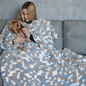 May include: A woman sitting on a sofa with a small dog, both wrapped in a grey, white and brown patterned blanket. The blanket is soft and fluffy, and the woman is smiling.