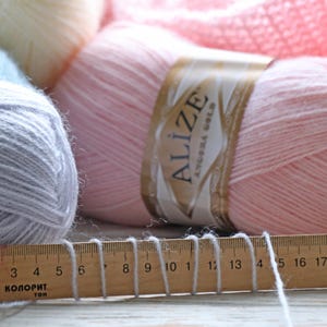 May include: A close-up of a pink ball of yarn with a white label that reads "Alize Angora Gold". The yarn is next to a wooden ruler with numbers 3 to 17.