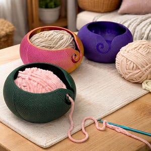 May include: Three yarn bowls in vibrant colors: a gradient of pink and yellow, a deep purple, and a forest green. Each bowl holds a different color of yarn, with a strand of pink yarn extending from the green bowl. A crochet hook is also visible.