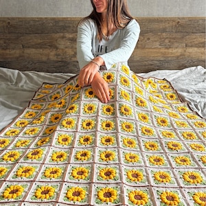 Puede incluir: Una manta de ganchillo hecha a mano con un patr&oacute;n repetido de girasoles. La manta tiene un fondo blanco con girasoles amarillos, centros marrones y hojas verdes. El borde es marr&oacute;n. La manta se muestra en una cama.