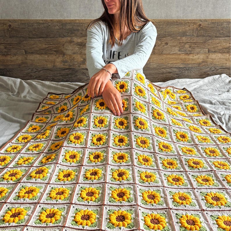 Puede incluir: Una manta de ganchillo hecha a mano con un patr&oacute;n repetido de girasoles. La manta tiene un fondo blanco con girasoles amarillos, centros marrones y hojas verdes. El borde es marr&oacute;n. La manta se muestra en una cama.