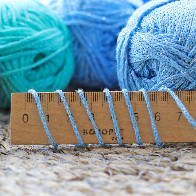 May include: A wooden ruler with blue yarn wrapped around it. The ruler has numbers 0 through 7 marked on it. The yarn is light blue and has a slight shimmer.