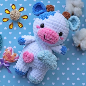 May include: A handmade crochet cow plushie in white, blue, pink, and brown. The cow has a pink nose, a heart-shaped patch, and blue accents on its hooves and ears. It is set against a blue background with white hearts.