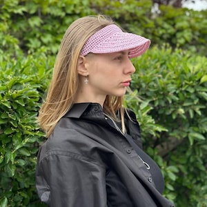 May include: A light pink crocheted visor with a wide brim. The visor is worn on the head. The background is a green leafy bush. The person is wearing a black shirt.