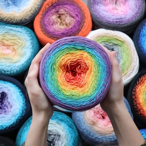 May include: A rainbow ombre yarn ball is held in someone's hands. The yarn ball is surrounded by other yarn balls in various colours, including blue, purple, pink, and orange.