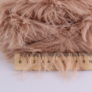 May include: Close-up of a swatch of light brown faux fur with long, soft fibers. A wooden ruler with black markings is placed across the fur, showing measurements in inches. The fur has a fluffy, textured appearance.