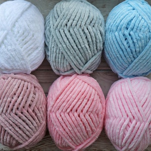 K&ouml;nnte beinhalten: Sechs Kn&auml;uel Garn in verschiedenen Farben: Wei&szlig;, Grau, Hellblau, Rosa und ein dunklerer Rosaton. Das Garn ist flauschig und texturiert.