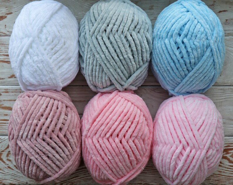 K&ouml;nnte beinhalten: Sechs Kn&auml;uel Garn in verschiedenen Farben: Wei&szlig;, Grau, Hellblau, Rosa und ein dunklerer Rosaton. Das Garn ist flauschig und texturiert.