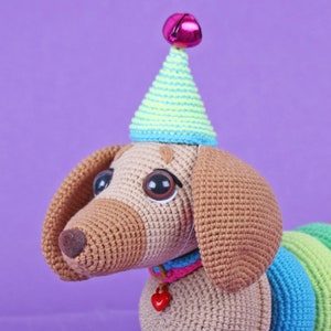May include: A crocheted dachshund wearing a party hat with a pink bell on top. The dachshund has brown fur, big brown eyes, and a rainbow-colored tail.
