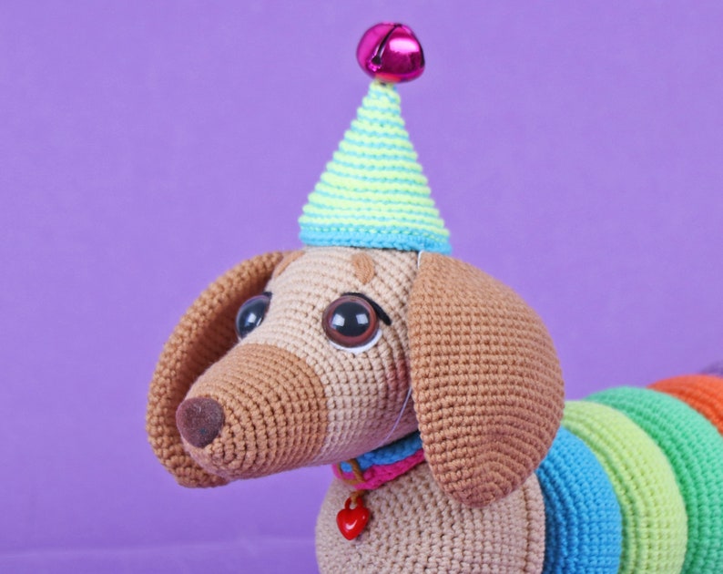 May include: A crocheted dachshund wearing a party hat with a pink bell on top. The dachshund has brown fur, big brown eyes, and a rainbow-colored tail.