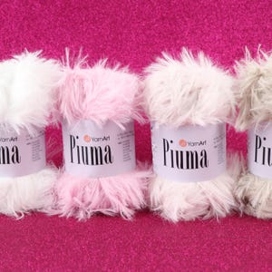 May include: Close-up of four spools of YarnArt Piuma yarn in white, pink, and gray. The yarn has a fluffy, feathery texture. The spools are on a bright pink, textured surface. The brand name "Piuma" is printed on the label.