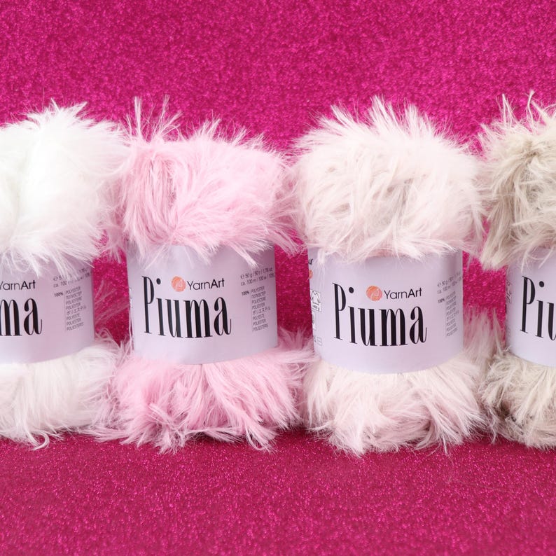 May include: Close-up of four spools of YarnArt Piuma yarn in white, pink, and gray. The yarn has a fluffy, feathery texture. The spools are on a bright pink, textured surface. The brand name "Piuma" is printed on the label.