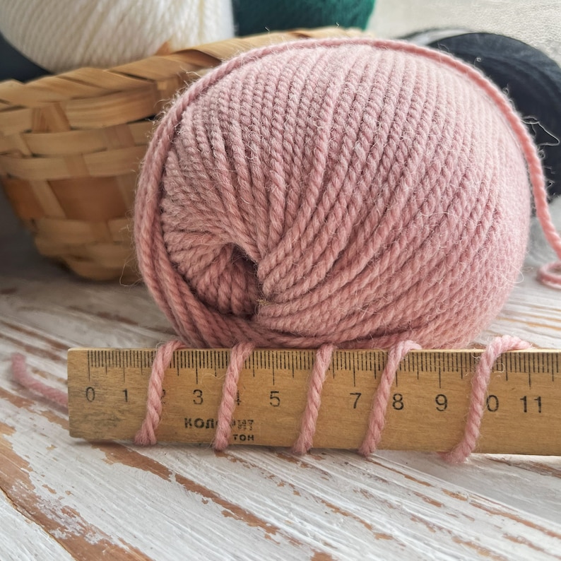 May include: A ball of pale pink yarn with a wooden ruler measuring 11 inches long. The yarn is wrapped around the ruler, with the ends of the yarn extending beyond the ruler.